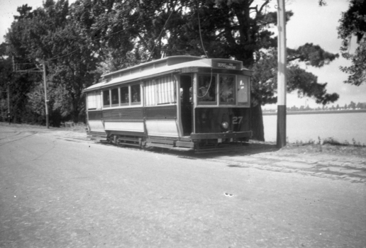 A two-colored vintage tram numbered 27 is stopped on tracks next to a road with a body of water and trees in the background.