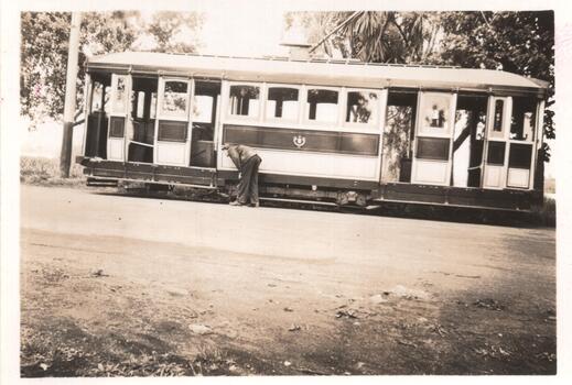 A man in work clothes bends down to tend to the tracks of a vintage tramcar. The tram is open-sided, revealing multiple windows and doors.