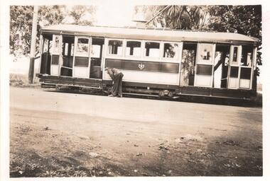 A man in work clothes bends down to tend to the tracks of a vintage tramcar. The tram is open-sided, revealing multiple windows and doors.