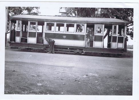A man in work clothes bends down to tend to the tracks of a vintage tramcar. The tram is open-sided, revealing multiple windows and doors.