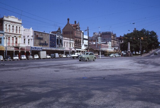 Street scene of a town with historic buildings, shops, trams, cars, and a monument, under a clear blue sky.
