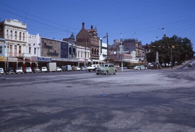 Street scene of a town with historic buildings, shops, trams, cars, and a monument, under a clear blue sky.