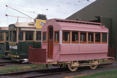 A pink vintage tram with wooden-framed windows sits on tracks next to a green and cream tram.  The tram is painted pink and is being rebuilt.
