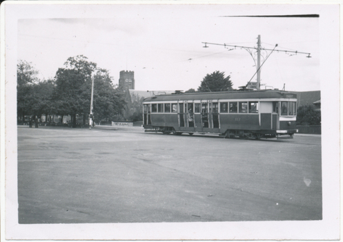  vintage tram with the number 117 is stopped on a street in front of a brick building and trees. The tram has large windows and multiple doors along its side.
