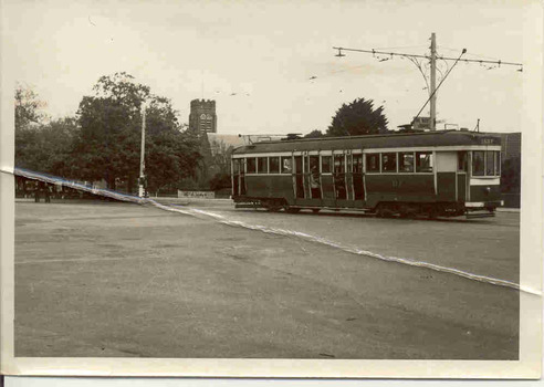  vintage tram with the number 117 is stopped on a street in front of a brick building and trees. The tram has large windows and multiple doors along its side.