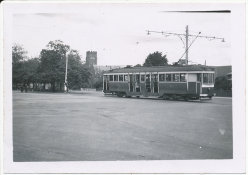  vintage tram with the number 117 is stopped on a street in front of a brick building and trees. The tram has large windows and multiple doors along its side.