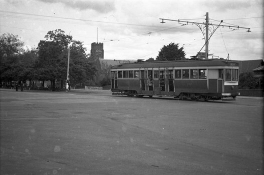  vintage tram with the number 117 is stopped on a street in front of a brick building and trees. The tram has large windows and multiple doors along its side.