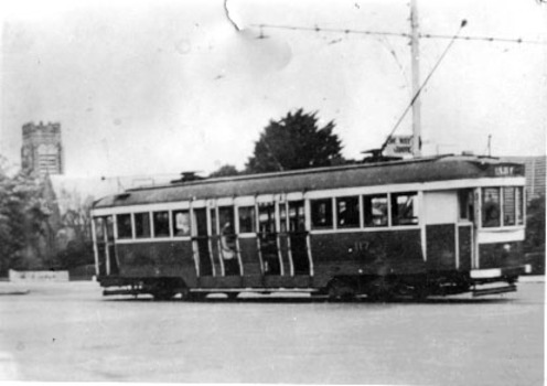  vintage tram with the number 117 is stopped on a street in front of a brick building and trees. The tram has large windows and multiple doors along its side.