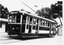 Shows tram numbered 29. The tram has several windows and appears to be stopped at a depot, with a building to the left and trees in the background.
