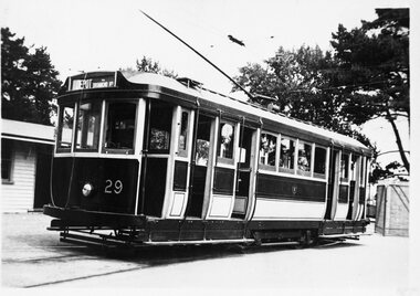 Shows tram numbered 29. The tram has several windows and appears to be stopped at a depot, with a building to the left and trees in the background.
