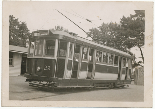 Shows tram numbered 29. The tram has several windows and appears to be stopped at a depot, with a building to the left and trees in the background.
