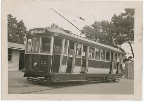 Shows tram numbered 29. The tram has several windows and appears to be stopped at a depot, with a building to the left and trees in the background.