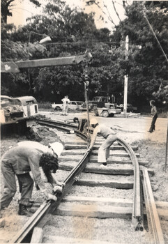Men in hard hats work on laying tram tracks, with a crane in the background. They are using tools to position the metal rails onto wooden sleepers.