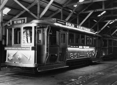 A number of vintage trams are parked indoors, with one bearing the text "VICTORIA ST", "GOLD CENTENARY" and "100 YEARS" and "1851 - 1951". The trams are in a large shed with wooden beams supporting the roof.