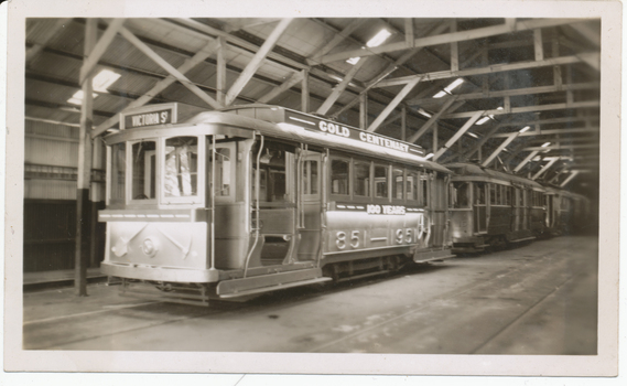 A number of vintage trams are parked indoors, with one bearing the text "VICTORIA ST", "GOLD CENTENARY" and "100 YEARS" and "1851 - 1951". The trams are in a large shed with wooden beams supporting the roof.