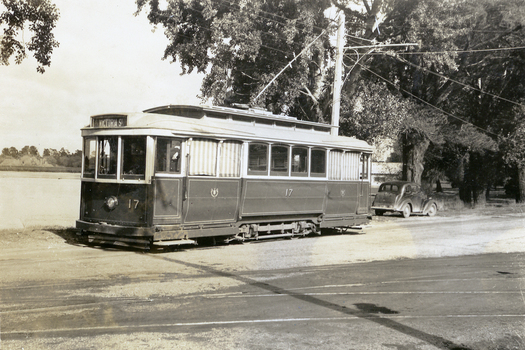 A vintage electric tram, number 17, with "Victoria St" displayed on its front, is parked on a track next to a body of water and trees. A vintage car is visible parked behind the tram.