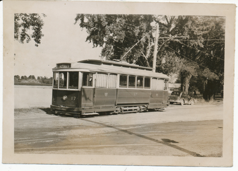 A vintage electric tram, number 17, with "Victoria St" displayed on its front, is parked on a track next to a body of water and trees. A vintage car is visible parked behind the tram.