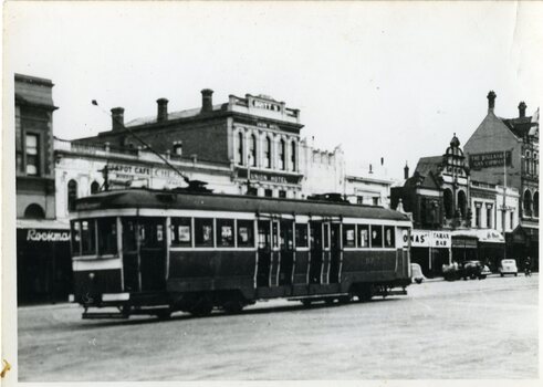 A historic tram car travels down a street lined with brick buildings, some featuring awnings and signs for businesses like "Union Hotel" and "Tarax Bar."