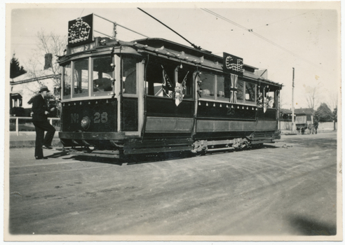 A decorated vintage tram numbered 28, with a driver in uniform stepping onto the platform, is parked on a street, adorned with Union Jack flags and ornate banners.