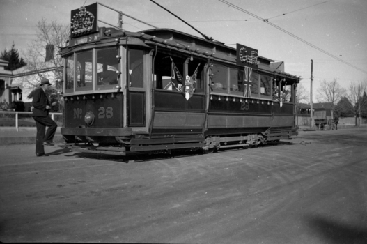 A decorated vintage tram numbered 28, with a driver in uniform stepping onto the platform, is parked on a street, adorned with Union Jack flags and ornate banners.