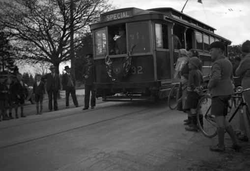A vintage tramcar labeled "SPECIAL" with the number 32 on its side, stopped on tracks beside a group of men and children, with a few bicycles nearby.
