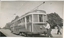 A tramcar labeled "GARDENS" and "STURT" is positioned on tracks next to a road with lush trees in the background. A man in a uniform and cap is holding a pole attached to the tram's overhead electrical system. Another person walks away in the background.