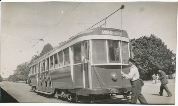 A tramcar labeled "GARDENS" and "STURT" is positioned on tracks next to a road with lush trees in the background. A man in a uniform and cap is holding a pole attached to the tram's overhead electrical system. Another person walks away in the background.