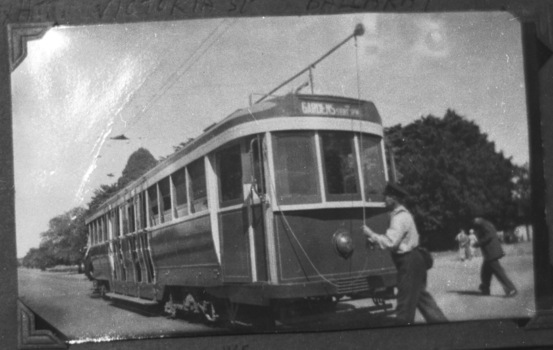 A tramcar labeled "GARDENS" and "STURT" is positioned on tracks next to a road with lush trees in the background. A man in a uniform and cap is holding a pole attached to the tram's overhead electrical system. Another person walks away in the background.