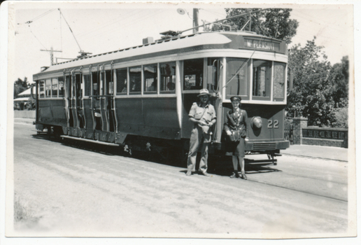 A vintage tram, numbered 22 and marked "MT. PLEASANT", stands on tracks next to a road. Two individuals, one in a khaki uniform and hat, and another, a lady conductor in a dark uniform with a cap, stand at the front of the tram. Trees and a brick fence are visible in the background.