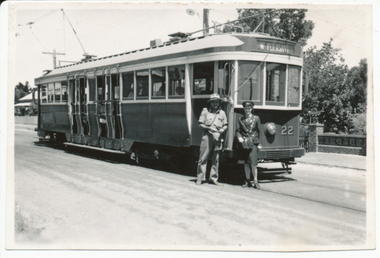 A vintage tram, numbered 22 and marked "MT. PLEASANT", stands on tracks next to a road. Two individuals, one in a khaki uniform and hat, and another, a lady conductor in a dark uniform with a cap, stand at the front of the tram. Trees and a brick fence are visible in the background.