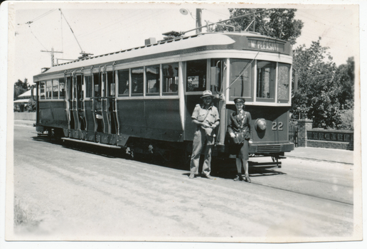 A vintage tram, numbered 22 and marked "MT. PLEASANT", stands on tracks next to a road. Two individuals, one in a khaki uniform and hat, and another, a lady conductor in a dark uniform with a cap, stand at the front of the tram. Trees and a brick fence are visible in the background.