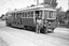 A vintage tram, numbered 22 and marked "MT. PLEASANT", stands on tracks next to a road. Two individuals, one in a khaki uniform and hat, and another, a lady conductor in a dark uniform with a cap, stand at the front of the tram. Trees and a brick fence are visible in the background.