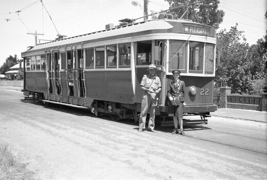 A vintage tram, numbered 22 and marked "MT. PLEASANT", stands on tracks next to a road. Two individuals, one in a khaki uniform and hat, and another, a lady conductor in a dark uniform with a cap, stand at the front of the tram. Trees and a brick fence are visible in the background.