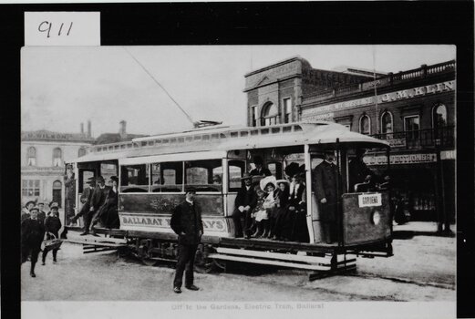 Black and white historical image of an open-air electric tram filled with passengers, with buildings lining the street and a sign on the tram reading "Gardens" and "Ballarat Tramways".