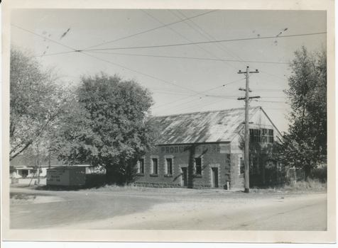 A brick building with a corrugated metal roof and the word "PRODUCE" visible on its facade, situated next to a large tree and a utility pole with overhead wires.
