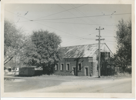 A brick building with a corrugated metal roof and the word "PRODUCE" visible on its facade, situated next to a large tree and a utility pole with overhead wires.