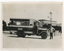 A vintage bus, labeled "BALLARAT TRAMWAYS" and "MT PLEASANT", is parked on a dirt road in front of a picket fence and a building with ornate trim. A man in a uniform stands by the driver's side of the bus, with his hand on the vehicle.
