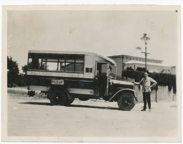 A vintage bus, labeled "BALLARAT TRAMWAYS" and "MT PLEASANT", is parked on a dirt road in front of a picket fence and a building with ornate trim. A man in a uniform stands by the driver's side of the bus, with his hand on the vehicle.
