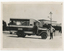 A vintage bus, labeled "BALLARAT TRAMWAYS" and "MT PLEASANT", is parked on a dirt road in front of a picket fence and a building with ornate trim. A man in a uniform stands by the driver's side of the bus, with his hand on the vehicle.