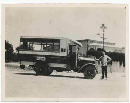 A vintage bus, labeled "BALLARAT TRAMWAYS" and "MT PLEASANT", is parked on a dirt road in front of a picket fence and a building with ornate trim. A man in a uniform stands by the driver's side of the bus, with his hand on the vehicle.