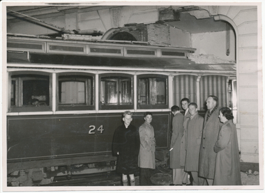Shows a damaged section of a building wall above a vintage tram labeled with the number 24. A group of individuals stands in front of the tram, appearing to observe the damage.