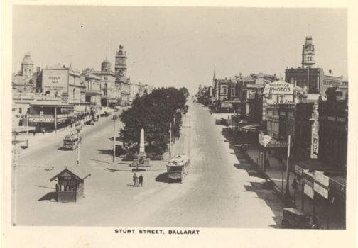 The street is lined with ornate Victorian-era buildings on both sides. A prominent clock tower stands tall on the left side of the street. A monument with an obelisk is situated in the center of the road, surrounded by trees. Trams are visible on the street.