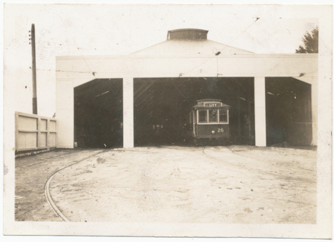 A vintage electric tram with number 26 and the word "CITY" on its front is positioned in the entrance of a large, open-sided tram shed.