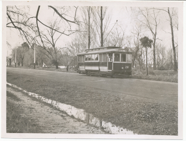 A vintage tram numbered 28 travels down a road lined with trees and a ditch.