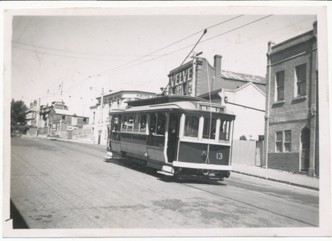 A vintage tram numbered 13 with "Victoria St" displayed on its side travels along a paved street lined with historic brick buildings.