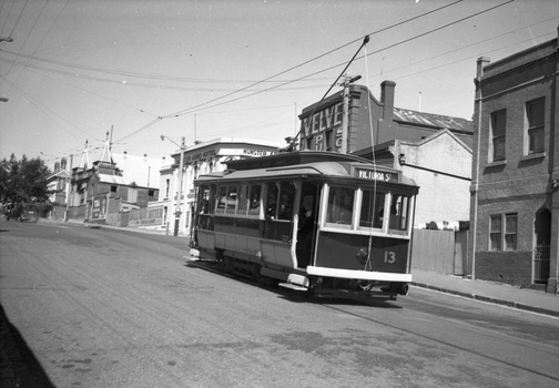 A vintage tram numbered 13 with "Victoria St" displayed on its side travels along a paved street lined with historic brick buildings.