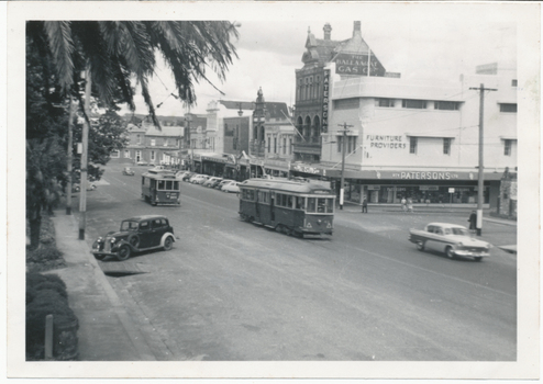 Shows a street scene in a town during the daytime. The street is populated by two vintage trams moving along tracks in the road, and several cars, including a dark sedan from the 1930s on the left and a white sedan from the 1950s on the right. Buildings line both sides of the street, with storefronts visible, including one with a sign reading "Patersons Furniture Providers." Palm fronds from a large tree arch over the left side of the image.