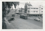 Shows a street scene in a town during the daytime. The street is populated by two vintage trams moving along tracks in the road, and several cars, including a dark sedan from the 1930s on the left and a white sedan from the 1950s on the right. Buildings line both sides of the street, with storefronts visible, including one with a sign reading "Patersons Furniture Providers." Palm fronds from a large tree arch over the left side of the image.