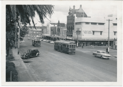 Shows a street scene in a town during the daytime. The street is populated by two vintage trams moving along tracks in the road, and several cars, including a dark sedan from the 1930s on the left and a white sedan from the 1950s on the right. Buildings line both sides of the street, with storefronts visible, including one with a sign reading "Patersons Furniture Providers." Palm fronds from a large tree arch over the left side of the image.