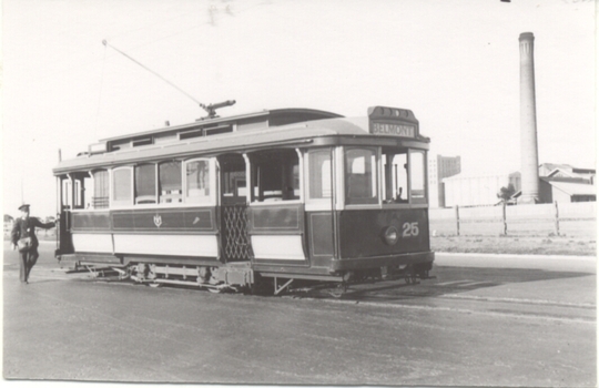 A vintage tram labeled "BELMONT" and "25" is stopped on a street next to a uniformed man. In the background, a tall industrial chimney and buildings are visible.